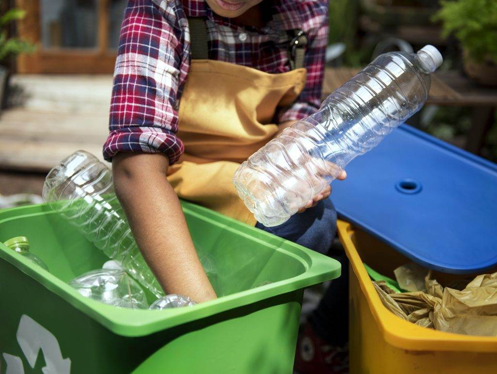 #PraCegoVer A imagem descreve uma cena de reciclagem consciente, onde uma pessoa, entre duas caçambas de lixo - uma verde e outra amarela - recolhe cuidadosamente garrafas PET.
